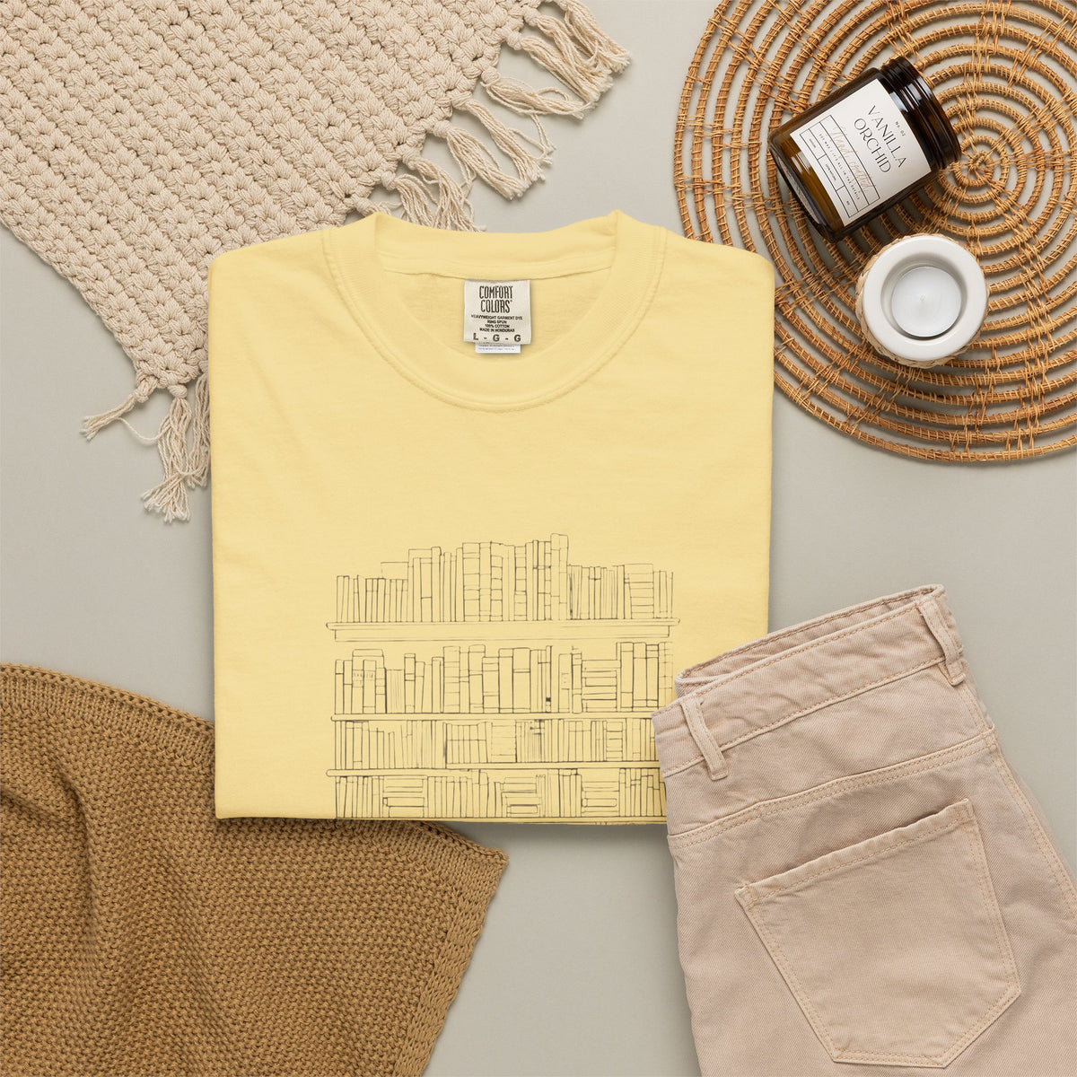 So Many Books So Little Shelf Space tee in Butter yellow, folded in a styled flat lay with a woven mat, candle and khaki shorts — bookshelf print on the front
