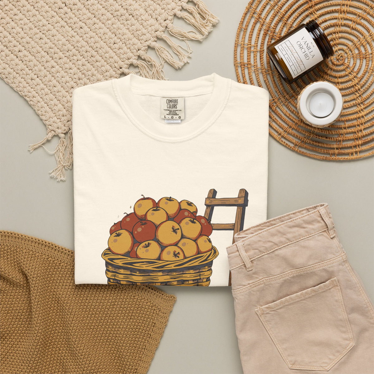 Peaked in the Orchard tee in Ivory, folded in a styled flat lay with a woven mat, candle and khaki shorts — apple basket print on the front