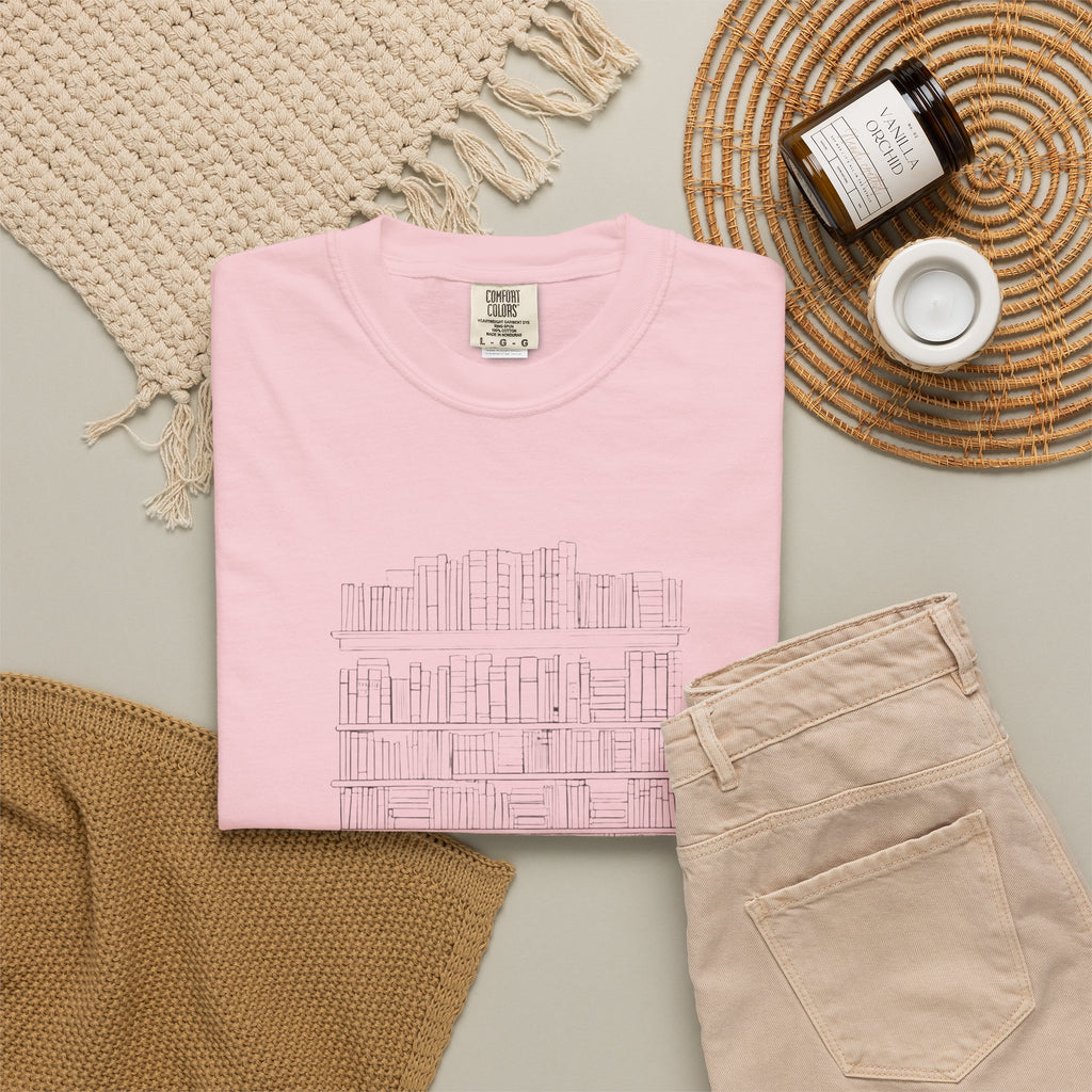 So Many Books So Little Shelf Space tee in Blossom pink, folded in a styled flat lay with a woven mat, candle and khaki shorts — bookshelf print on the front