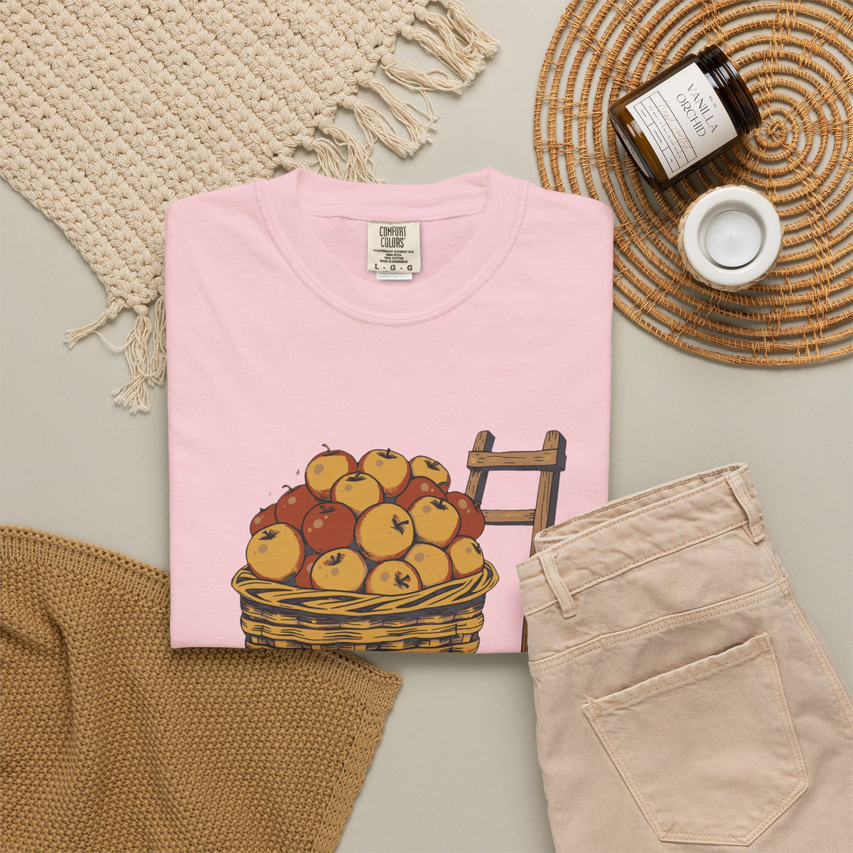 Peaked in the Orchard tee in Blossom pink, folded in a styled flat lay with a woven mat, candle and khaki shorts — apple basket print on the front
