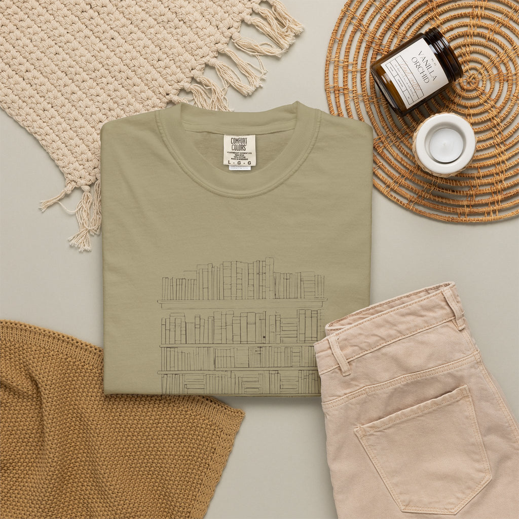 So Many Books So Little Shelf Space tee in Khaki, folded in a styled flat lay with a woven mat, candle and khaki shorts — bookshelf print on the front