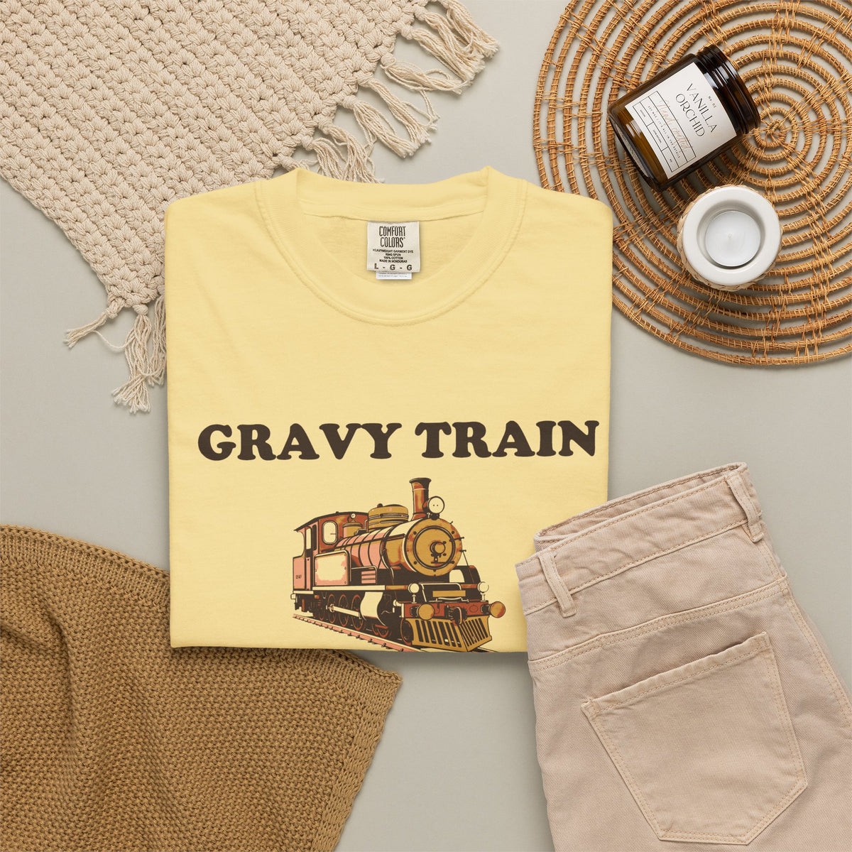 Gravy Train Conductor tee in Butter yellow, folded in a styled flat lay with a woven mat, candle and khaki shorts — locomotive print on the front