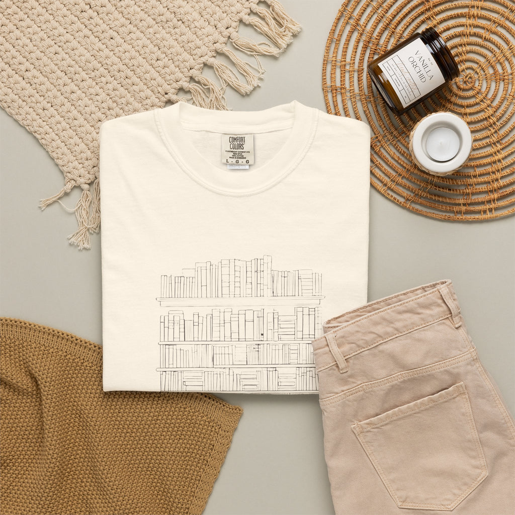 So Many Books So Little Shelf Space tee in Ivory, folded in a styled flat lay with a woven mat, candle and khaki shorts — bookshelf print on the front