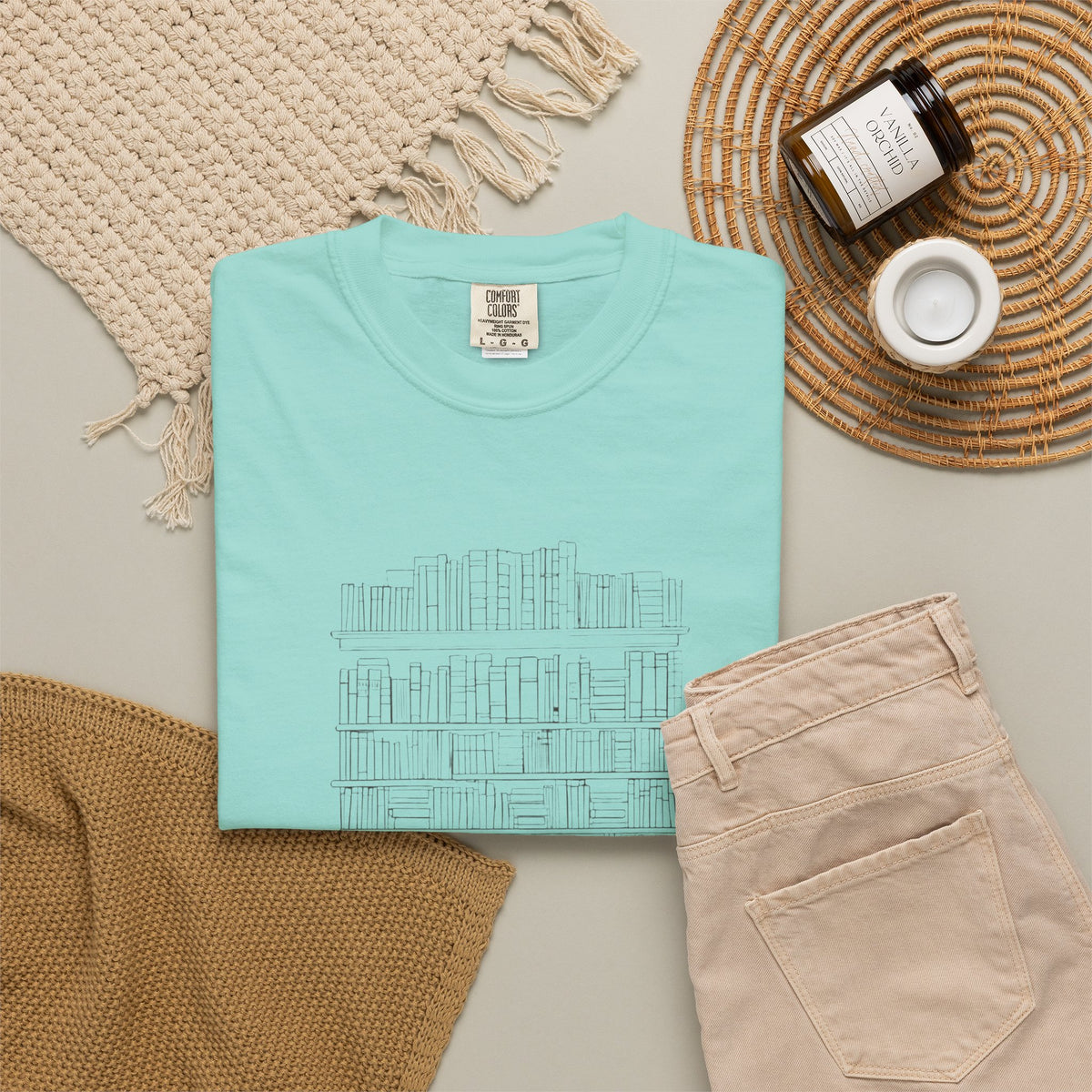 So Many Books So Little Shelf Space tee in Chalky Mint, folded in a styled flat lay with a woven mat, candle and khaki shorts — bookshelf print on the front