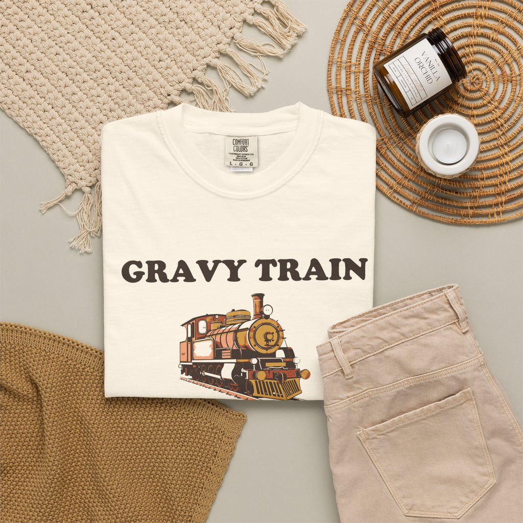 Gravy Train Conductor tee in Ivory, folded in a styled flat lay with a woven mat, candle and khaki shorts — locomotive print on the front