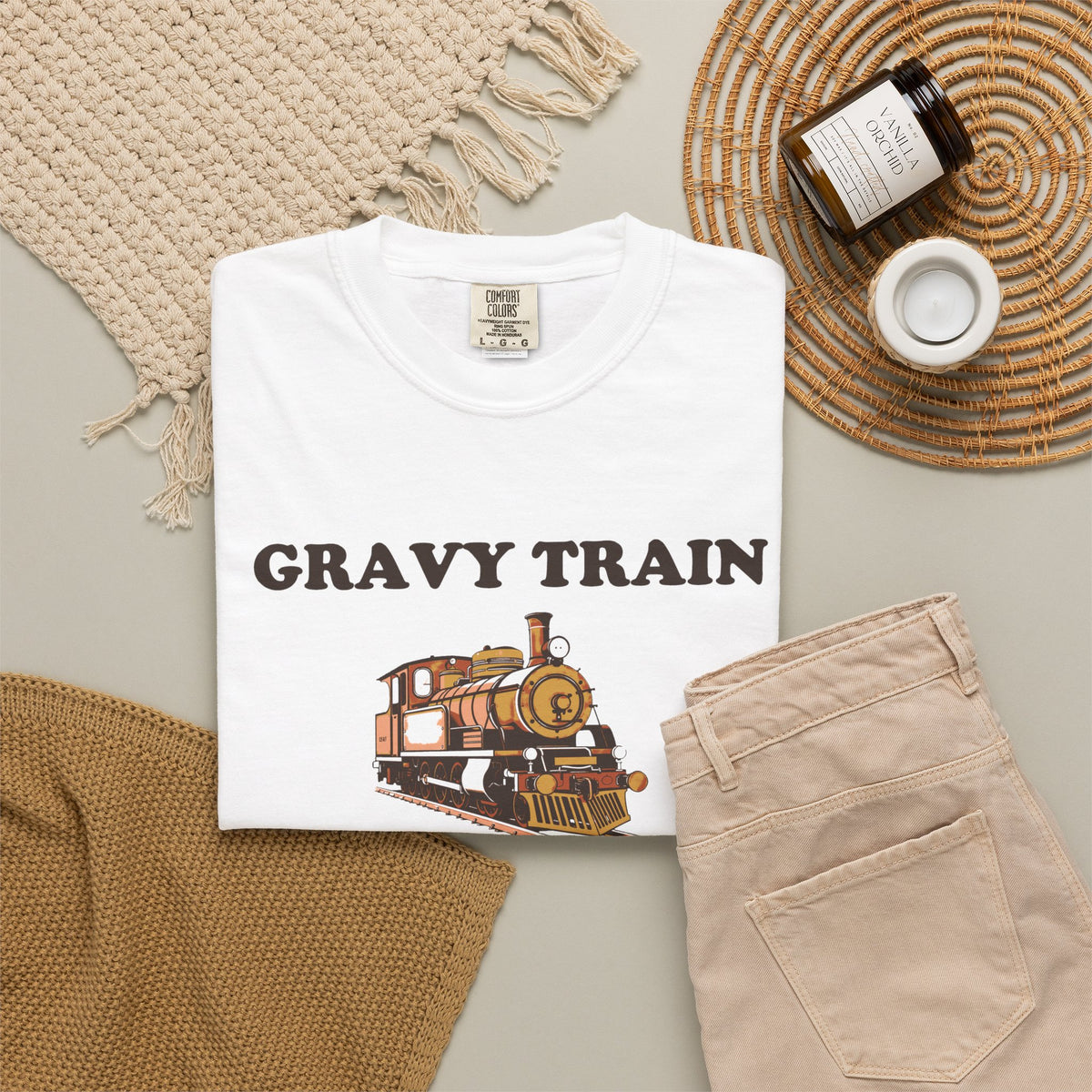 Gravy Train Conductor tee in White, folded in a styled flat lay with a woven mat, candle and khaki shorts — locomotive print on the front