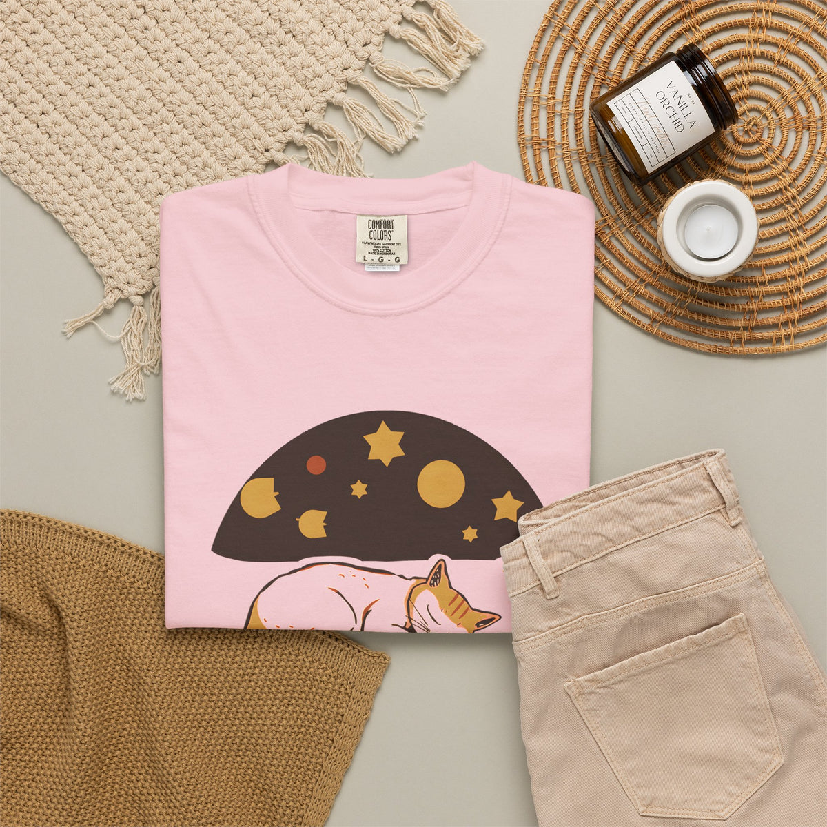 Lunch Break Read tee in Blossom pink, folded in a styled flat lay with a woven mat, candle and khaki shorts — sleeping cat print on the front