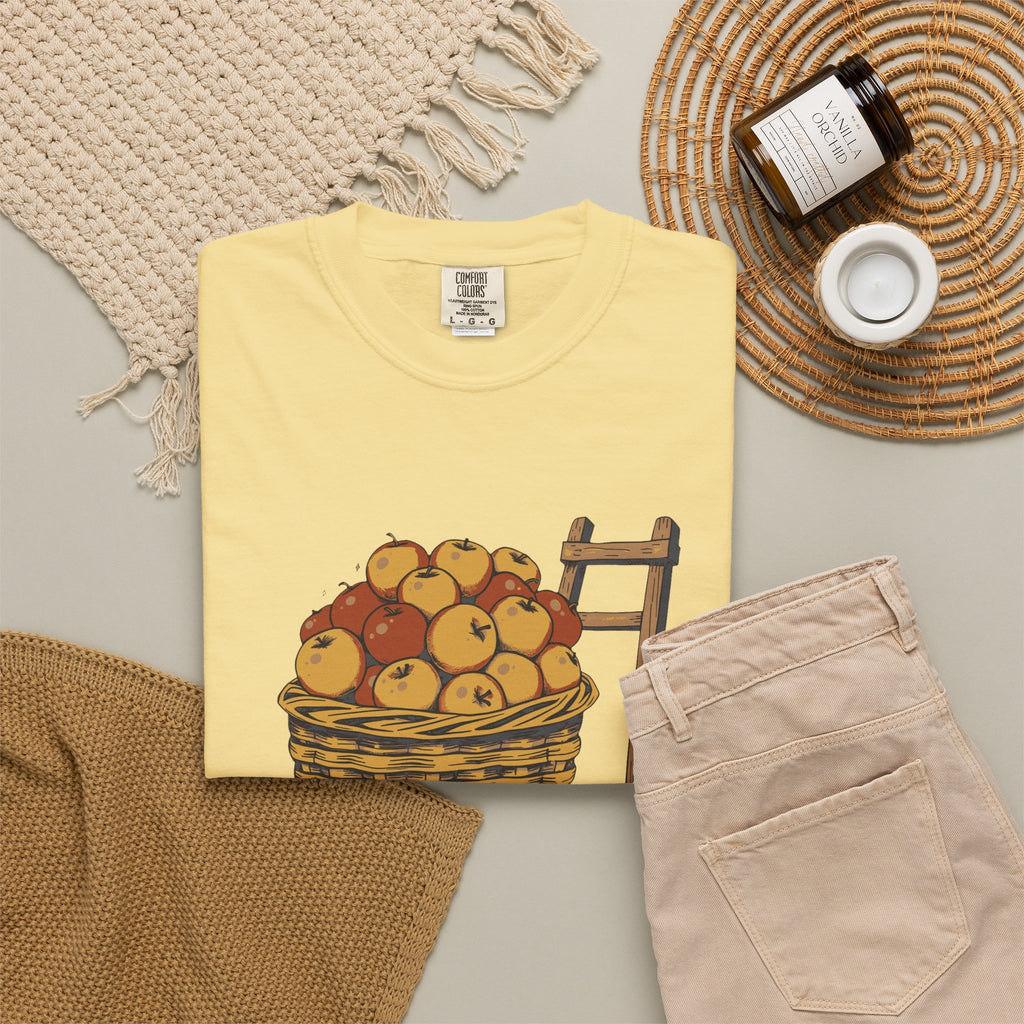 Peaked in the Orchard tee in Butter yellow, folded in a styled flat lay with a woven mat, candle and khaki shorts — apple basket print on the front