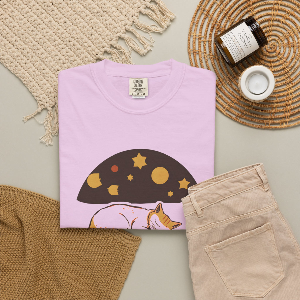 Lunch Break Read tee in Orchid, folded in a styled flat lay with a woven mat, candle and khaki shorts — sleeping cat print on the front
