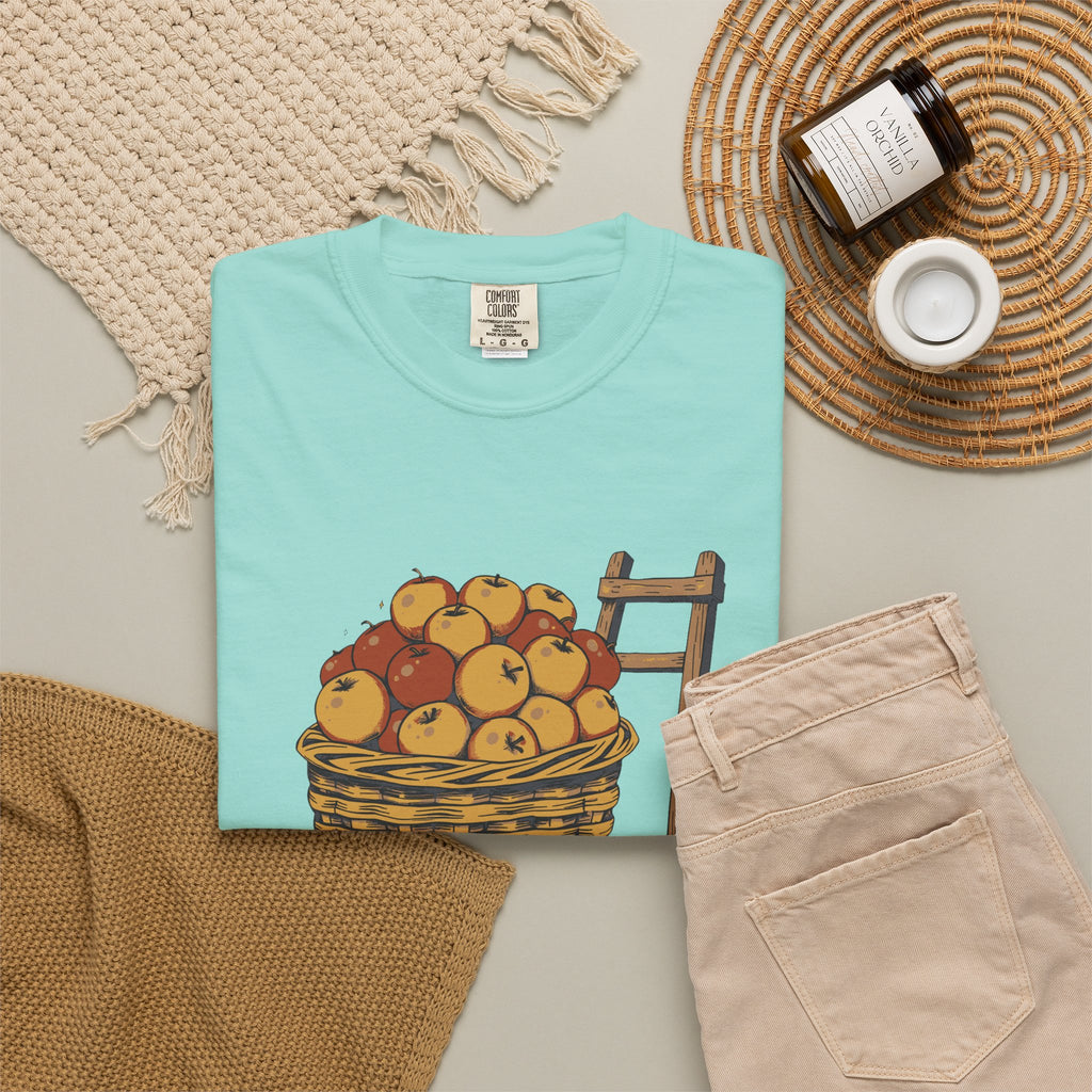 Peaked in the Orchard tee in Chalky Mint, folded in a styled flat lay with a woven mat, candle and khaki shorts — apple basket print on the front
